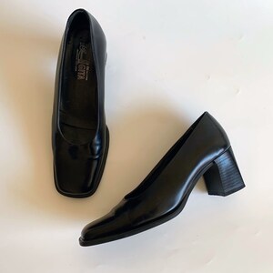 May include: A pair of black leather square-toe pumps. The shoes have a low block heel and a glossy finish. The brand name "Rossi Giva" is visible on the insole. The shoes are displayed on a white surface.