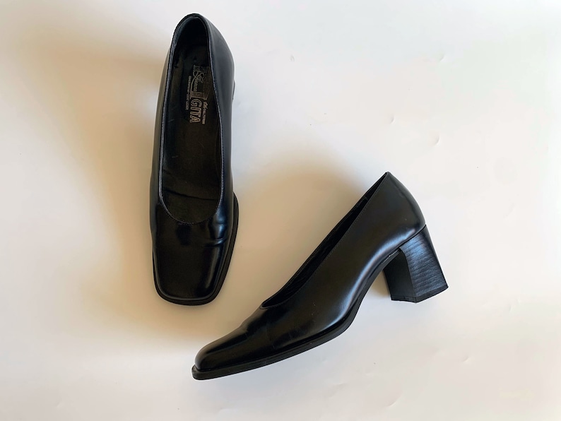 May include: A pair of black leather square-toe pumps. The shoes have a low block heel and a glossy finish. The brand name "Rossi Giva" is visible on the insole. The shoes are displayed on a white surface.