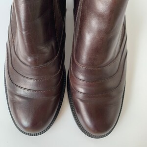 K&ouml;nnte beinhalten: Ein Paar braune Lederstiefeletten mit abgerundeter Spitze und einem dekorativen Nahtdetail. Die Stiefeletten haben ein glattes Lederfinish und einen leicht erh&ouml;hten Absatz.