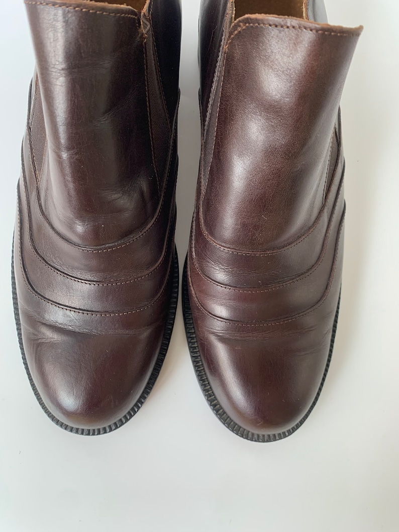 K&ouml;nnte beinhalten: Ein Paar braune Lederstiefeletten mit abgerundeter Spitze und einem dekorativen Nahtdetail. Die Stiefeletten haben ein glattes Lederfinish und einen leicht erh&ouml;hten Absatz.