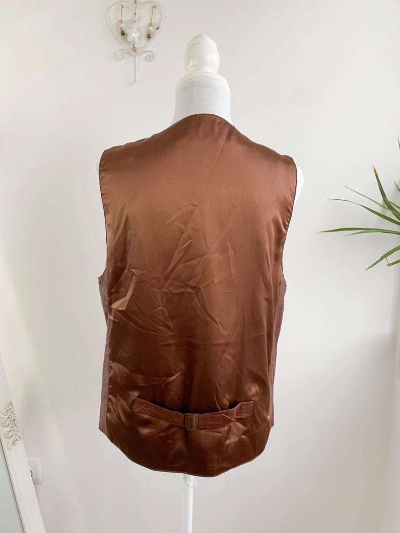 May include: Back view of a brown vest on a mannequin. The vest has a satin-like sheen and a small belt detail at the waist. The vest is displayed against a plain white background.