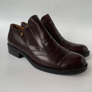 K&ouml;nnte beinhalten: Ein Paar braune Lederstiefeletten mit Schlupfdesign. Die Stiefeletten haben eine abgerundete Spitze und einen niedrigen Blockabsatz.