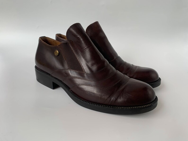 K&ouml;nnte beinhalten: Ein Paar braune Lederstiefeletten mit Schlupfdesign. Die Stiefeletten haben eine abgerundete Spitze und einen niedrigen Blockabsatz.