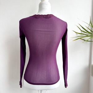May include: A long-sleeved, deep purple shirt displayed on a mannequin. The shirt features a delicate lace trim around the neckline and cuffs. The fabric appears lightweight and slightly sheer, with a fitted silhouette.