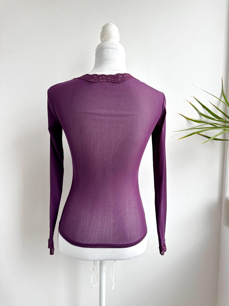 May include: A long-sleeved, deep purple shirt displayed on a mannequin. The shirt features a delicate lace trim around the neckline and cuffs. The fabric appears lightweight and slightly sheer, with a fitted silhouette.