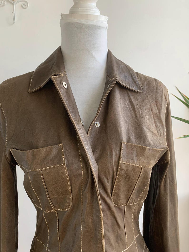 Op de afbeelding: Een bruine leren jas met een kraag en twee voorzakken. De jas heeft zichtbare stiksels in een lichtere bruine kleur. De jas met lange mouwen wordt op een mannequin gepresenteerd.