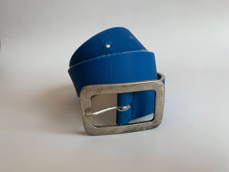 Pu&ograve; includere: Una cintura in pelle blu con una fibbia argentata. La cintura &egrave; piegata a met&agrave; e la fibbia &egrave; sul lato destro dell'immagine.