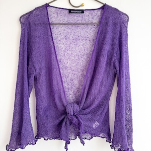 May include: A sheer, purple, long-sleeved bolero sweater with a tie closure at the waist. The sweater has a delicate, open weave and a scalloped edge on the sleeves. The label reads "NATURALLY".