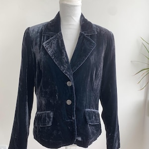 May include: A dark navy blue velvet blazer with a notched lapel, two front pockets, and two decorative buttons. The blazer is displayed on a white mannequin and has long sleeves. The fabric has a soft, textured appearance.