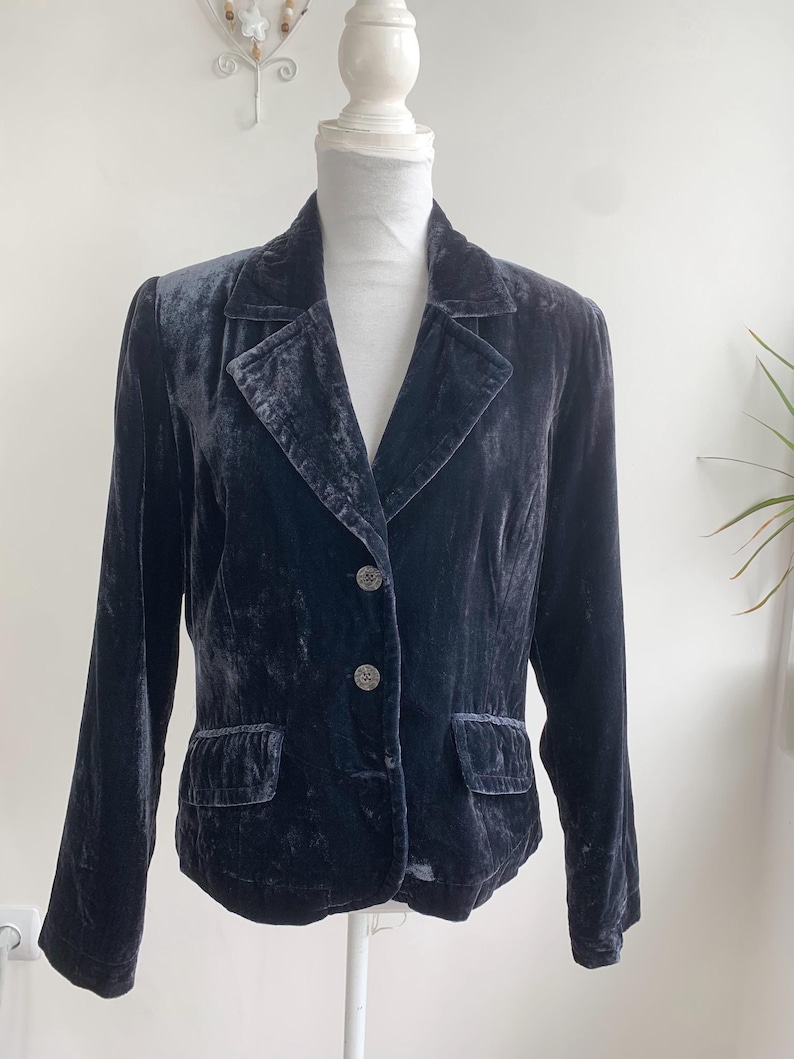 May include: A dark navy blue velvet blazer with a notched lapel, two front pockets, and two decorative buttons. The blazer is displayed on a white mannequin and has long sleeves. The fabric has a soft, textured appearance.