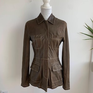 Op de afbeelding: Bruine leren jas met een kraag en vier voorzakken. De jas heeft lange mouwen en wordt op een mannequin gepresenteerd. De stiksels zijn lichtbruin van kleur, wat een contrast cre&euml;ert met het donkerdere leer.