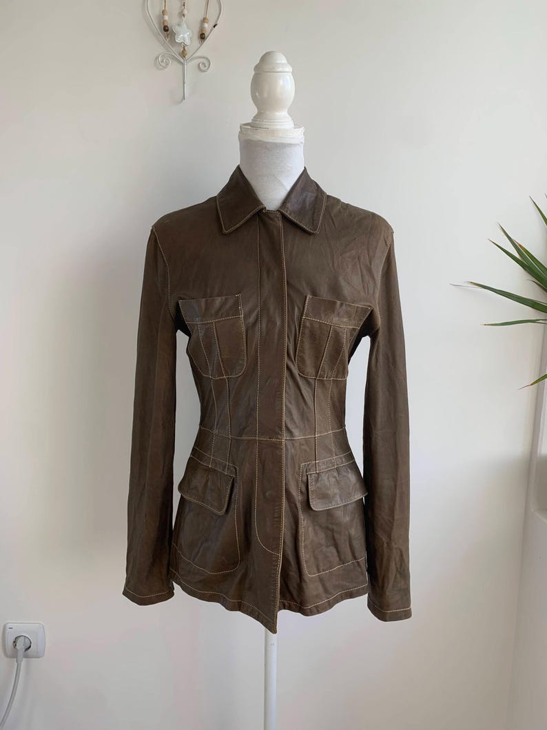 Op de afbeelding: Bruine leren jas met een kraag en vier voorzakken. De jas heeft lange mouwen en wordt op een mannequin gepresenteerd. De stiksels zijn lichtbruin van kleur, wat een contrast cre&euml;ert met het donkerdere leer.
