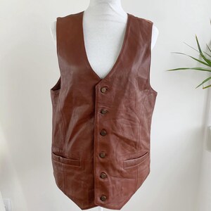 Puede incluir: Un chaleco de cuero marrón con cuello en V y cierre de botones. El chaleco tiene dos bolsillos delanteros y se exhibe en un maniquí blanco. El chaleco es de color marrón medio.
