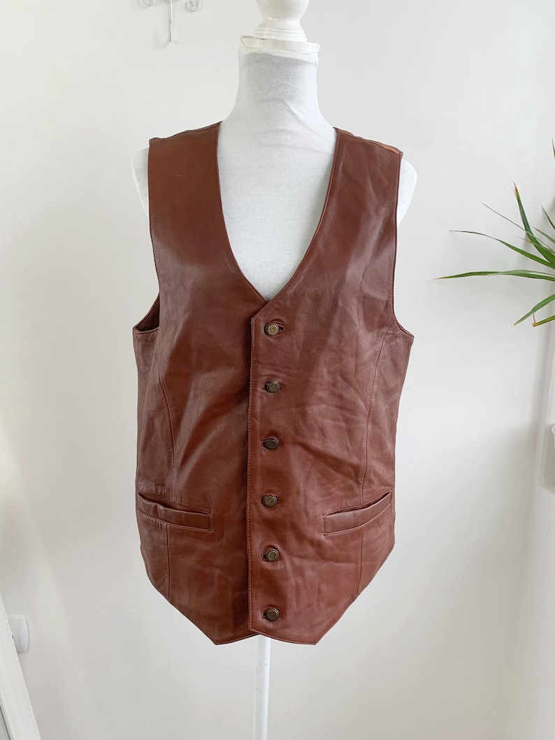 May include: A brown leather vest with a V-neck and button closure. The vest has two front pockets and is displayed on a white mannequin. The vest is a medium brown color.