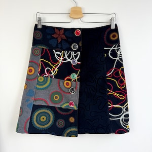May include: A patchwork mini skirt with a variety of patterns and colors. The skirt features a mix of fabrics, including a black fabric with a swirl pattern, and panels with colorful circles and floral designs. It has a button closure and is hanging on a hanger.