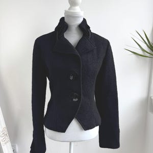 May include: A black wool jacket with a high collar and three large buttons. The jacket has long sleeves and a tailored fit, displayed on a white mannequin. The background is a plain white wall with a hint of a plant.