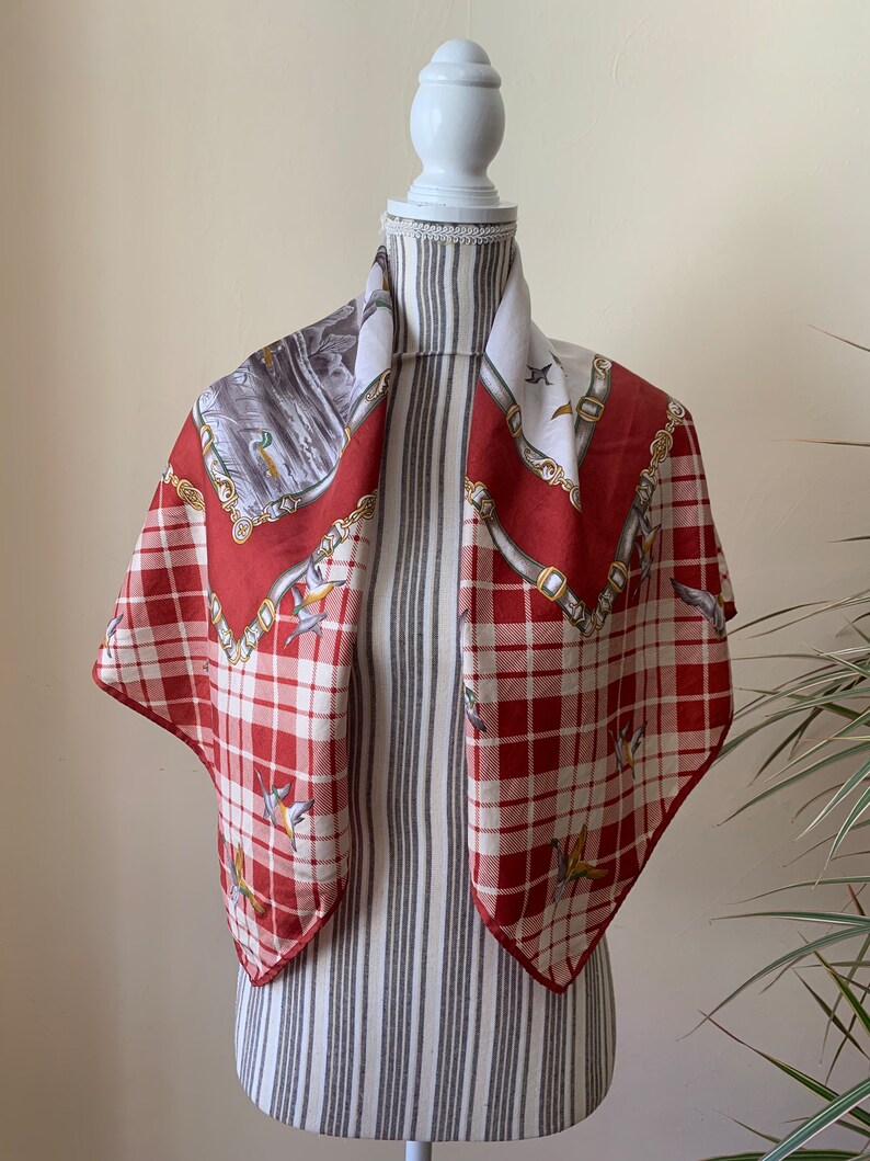 May include: A red and white plaid silk scarf with a gold chain and bird design. The scarf is displayed on a white mannequin.