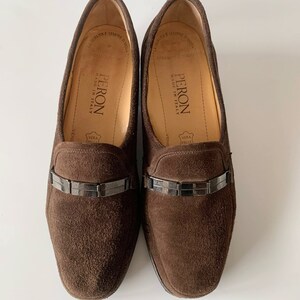 May include: A pair of brown suede loafers with a silver buckle detail. The loafers are made in Italy and have the brand name "PERON" on the insole.