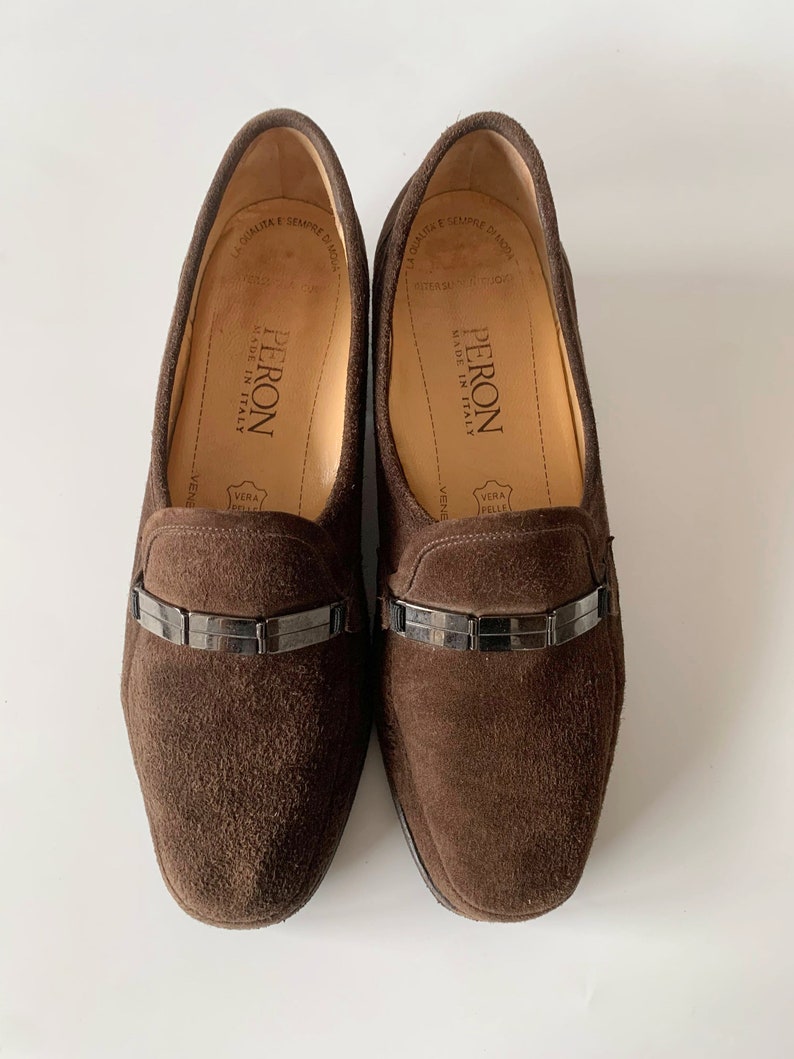 May include: A pair of brown suede loafers with a silver buckle detail. The loafers are made in Italy and have the brand name "PERON" on the insole.