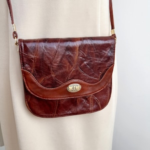 Op de afbeelding: Een bruine leren crossbodytas met een bladachtig patroon op de voorklep. De tas heeft een gebogen, donkerbruin accent en een goudkleurige metalen sluiting met de tekst "FASHION BAGS". De riem is dun en bruin.