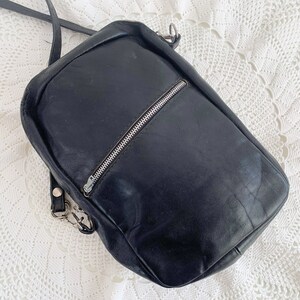 Pu&ograve; includere: Una borsa a tracolla in pelle nera con una cerniera argentata e una tracolla argentata. La borsa &egrave; su una superficie di uncinetto bianca.