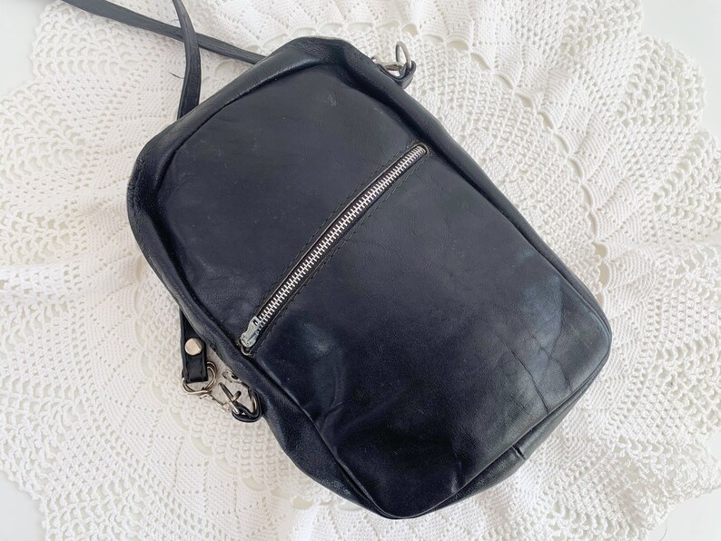 Pu&ograve; includere: Una borsa a tracolla in pelle nera con una cerniera argentata e una tracolla argentata. La borsa &egrave; su una superficie di uncinetto bianca.