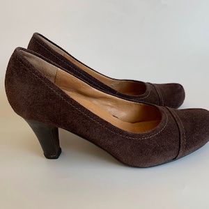 May include: A pair of brown suede heeled pumps. The shoes have a pointed toe and a closed back.