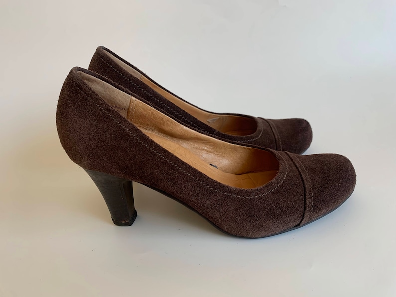 May include: A pair of brown suede heeled pumps. The shoes have a pointed toe and a closed back.