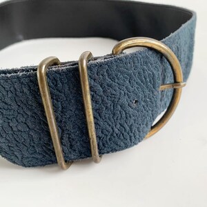 May include: A navy blue suede belt with a gold-coloured buckle and two gold-coloured loops. The belt has a textured surface and a black interior. The belt is a fashion accessory.