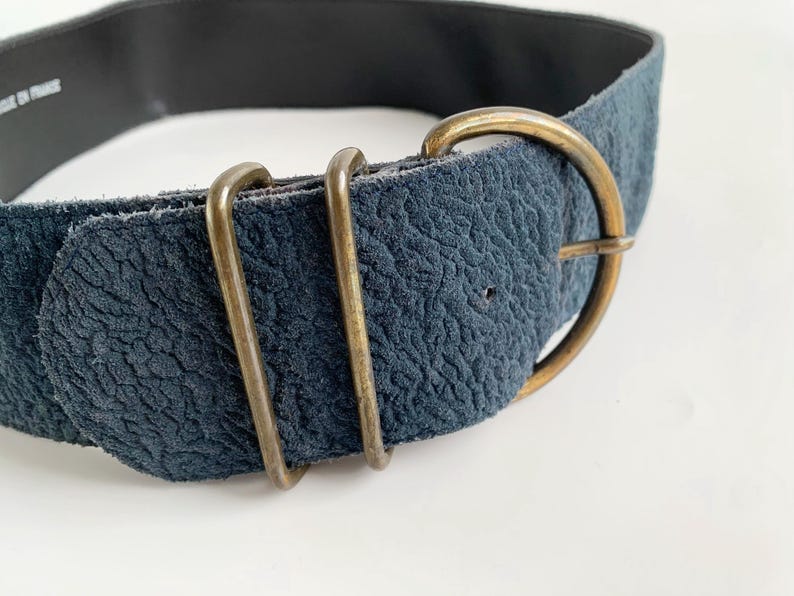 May include: A navy blue suede belt with a gold-coloured buckle and two gold-coloured loops. The belt has a textured surface and a black interior. The belt is a fashion accessory.