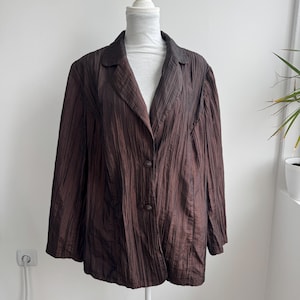 Pode incluir: Um blazer castanho escuro com um acabamento de tecido enrugado e texturizado. A peça apresenta uma gola de lapela com entalhe, mangas compridas e um fecho frontal fixado por dois botões castanhos escuros a condizer com um brilho subtil.