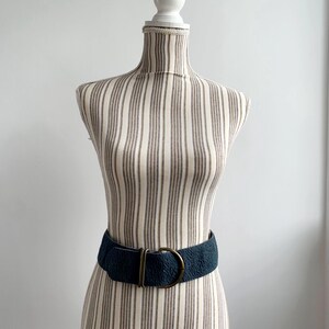 May include: A dark teal textured belt with a brass-coloured buckle is displayed on a striped mannequin. The belt is wide and cinches the waist. The mannequin is on a white background.
