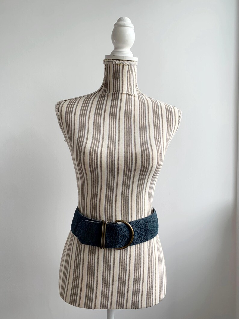 May include: A dark teal textured belt with a brass-coloured buckle is displayed on a striped mannequin. The belt is wide and cinches the waist. The mannequin is on a white background.