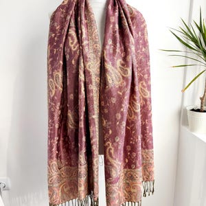May include: A burgundy paisley scarf with a cream and gold border and fringe. The scarf is draped over a white mannequin. The paisley pattern includes floral and teardrop shapes. The scarf is made of a soft, flowing material.