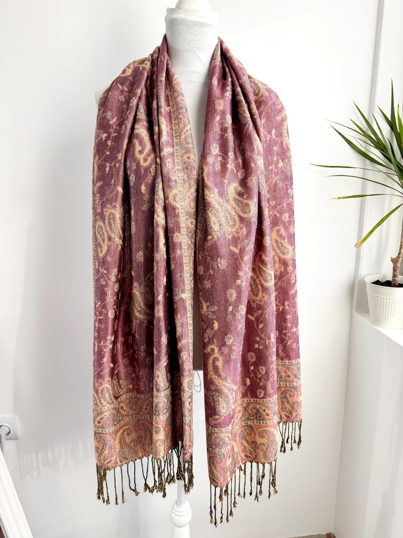 May include: A burgundy paisley scarf with a cream and gold border and fringe. The scarf is draped over a white mannequin. The paisley pattern includes floral and teardrop shapes. The scarf is made of a soft, flowing material.