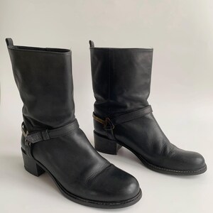 May include: A pair of black leather mid-calf boots featuring a rounded toe, a stacked block heel, and a decorative strap across the ankle secured with a metallic buckle detail. The boots have a pull tab at the top of the shaft and visible stitching.