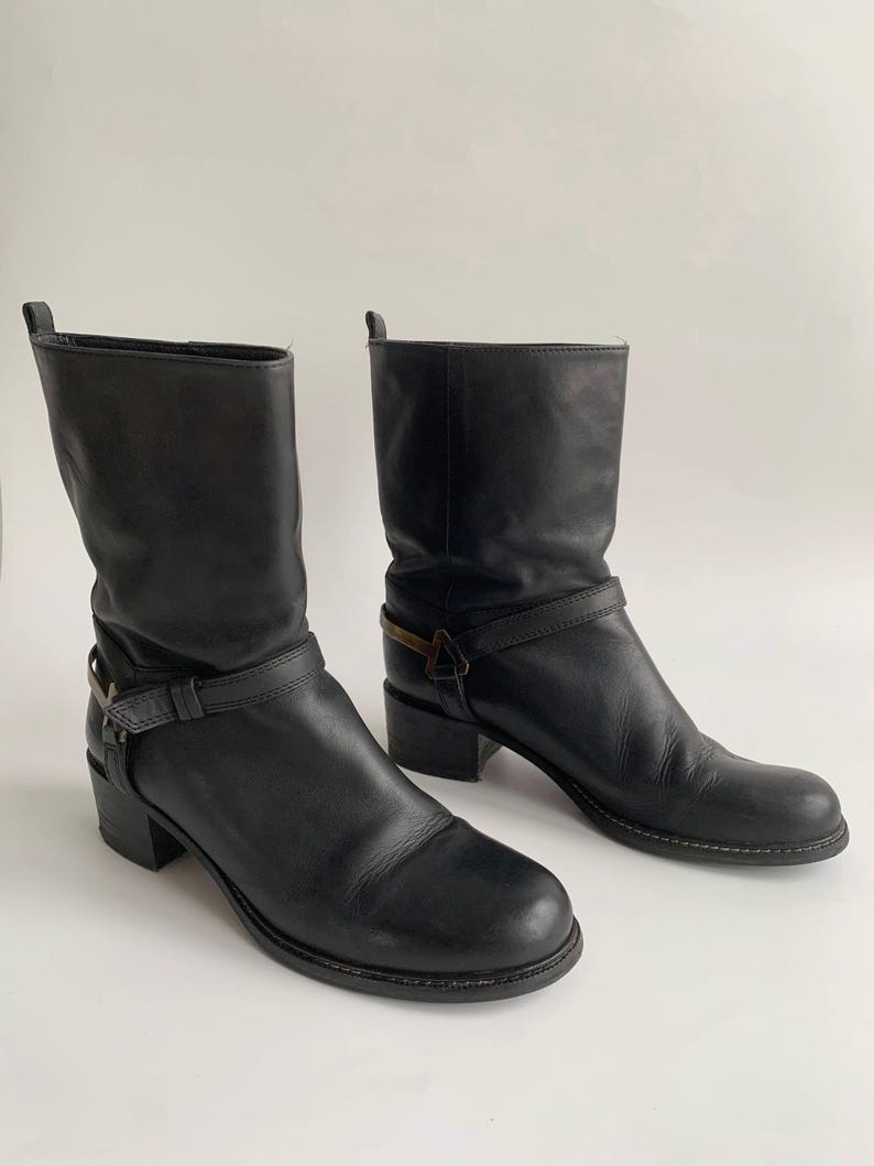 May include: A pair of black leather mid-calf boots featuring a rounded toe, a stacked block heel, and a decorative strap across the ankle secured with a metallic buckle detail. The boots have a pull tab at the top of the shaft and visible stitching.