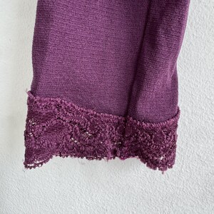 May include: Close-up of a section of a deep purple garment. The fabric has a textured appearance, and the edge is finished with a decorative, dark purple lace trim. The garment is against a white background.