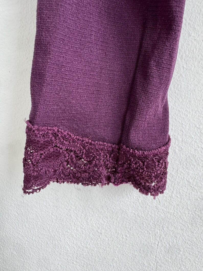 May include: Close-up of a section of a deep purple garment. The fabric has a textured appearance, and the edge is finished with a decorative, dark purple lace trim. The garment is against a white background.