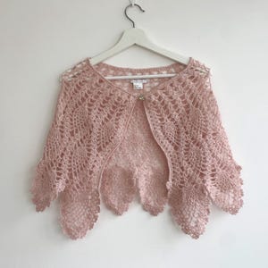 May include: A delicate pink crocheted shawl with a scalloped edge. The shawl is made of a lightweight, airy yarn and has a delicate, feminine look.