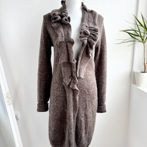 以下が含まれることがあります： フロントが開いた、茶色のロングカーディガン。前面と襟周りにフリルデザインがあります。袖口はリブ編みです。白いマネキンに着せています。柔らかく、質感のある素材で作られています。