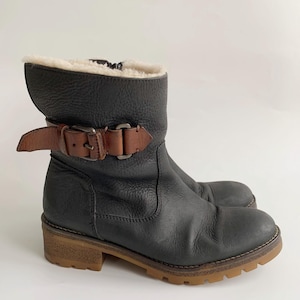 May include: A black leather ankle boot with a textured finish, featuring a brown leather strap with a metal buckle across the side. The boot has a cream-coloured faux fur lining at the top opening, a side zip, and a thick, lugged gum-coloured rubber sole.