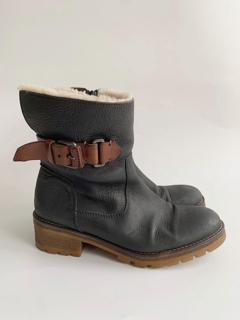 May include: A black leather ankle boot with a textured finish, featuring a brown leather strap with a metal buckle across the side. The boot has a cream-coloured faux fur lining at the top opening, a side zip, and a thick, lugged gum-coloured rubber sole.