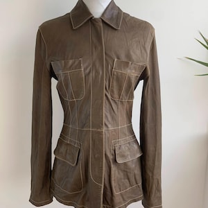 Op de afbeelding: Bruine leren jas met een kraag en vier voorzakken. De jas heeft lange mouwen en wordt op een mannequin getoond. De stiksels zijn lichtbruin van kleur, wat een contrast cre&euml;ert met de kleur van de jas.