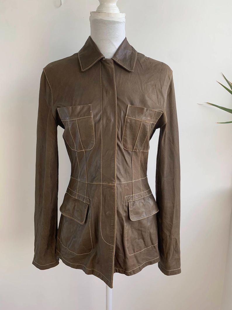Op de afbeelding: Bruine leren jas met een kraag en vier voorzakken. De jas heeft lange mouwen en wordt op een mannequin getoond. De stiksels zijn lichtbruin van kleur, wat een contrast cre&euml;ert met de kleur van de jas.