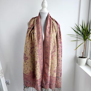 May include: A beige and burgundy paisley patterned scarf with fringe. The scarf is draped over a white mannequin. The scarf has a detailed paisley design in burgundy and beige tones. The fringe is a dark brown color.