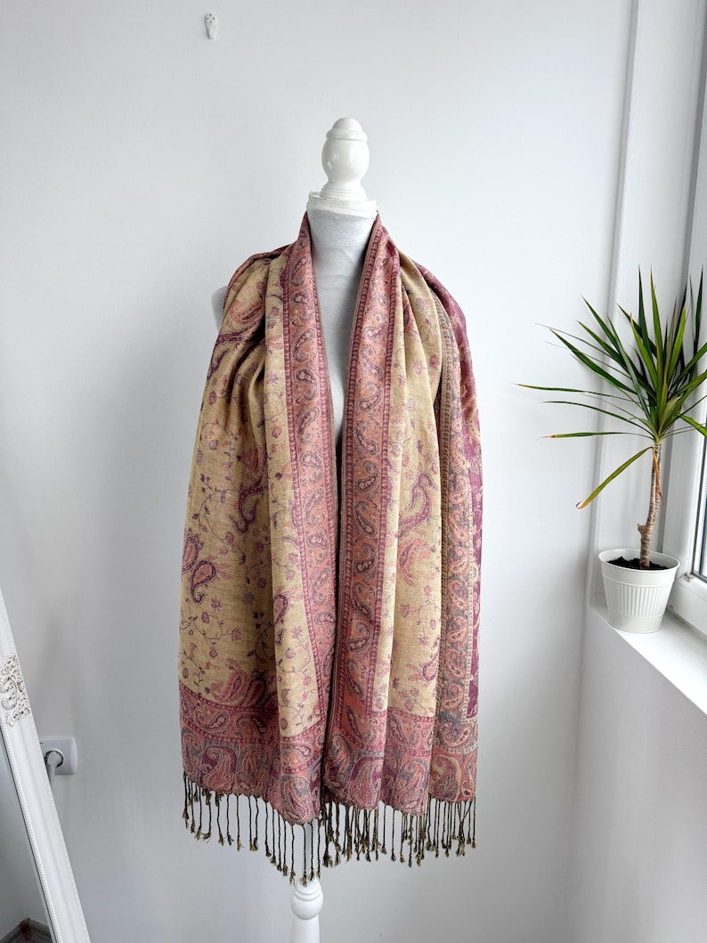 May include: A beige and burgundy paisley patterned scarf with fringe. The scarf is draped over a white mannequin. The scarf has a detailed paisley design in burgundy and beige tones. The fringe is a dark brown color.