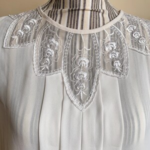 Puede incluir: Una blusa blanca con un delicado cuello de encaje con un dise&ntilde;o floral. La blusa tiene un frente plisado y una tela transparente.