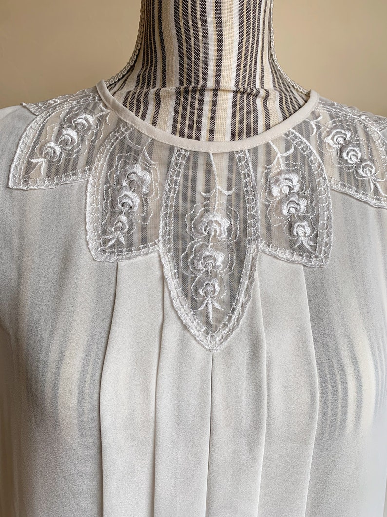 Puede incluir: Una blusa blanca con un delicado cuello de encaje con un dise&ntilde;o floral. La blusa tiene un frente plisado y una tela transparente.