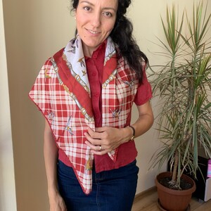 May include: A red and white plaid scarf with a white and gold patterned design. The scarf is tied around the neck of a person wearing a red button-down shirt and blue jeans.